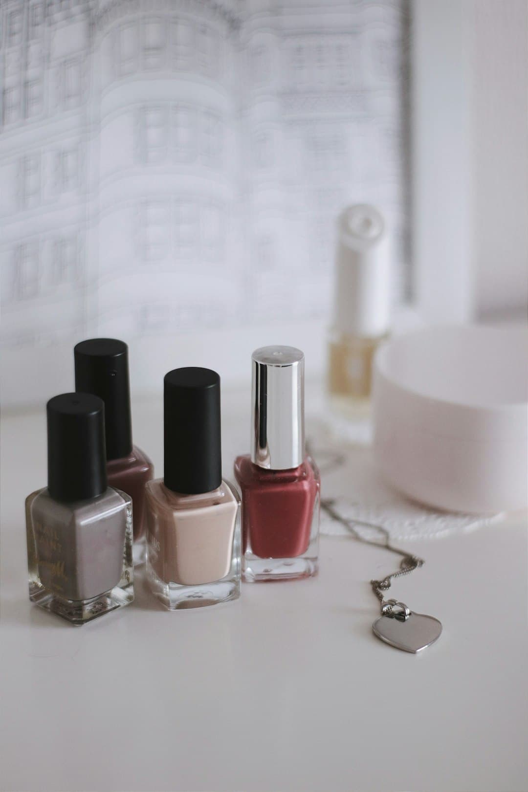 A still life of lacquer bottles — grey, nude and dusty-red glass with a silver cap — on a warm shelf.